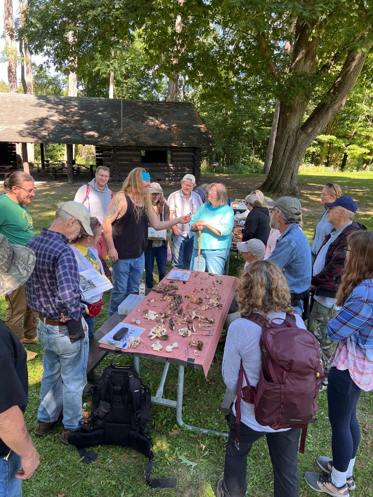 RAMA participants gathered around a picnic table covered with mushrooms, examining and discussing their finds beside a wooded pavilion.