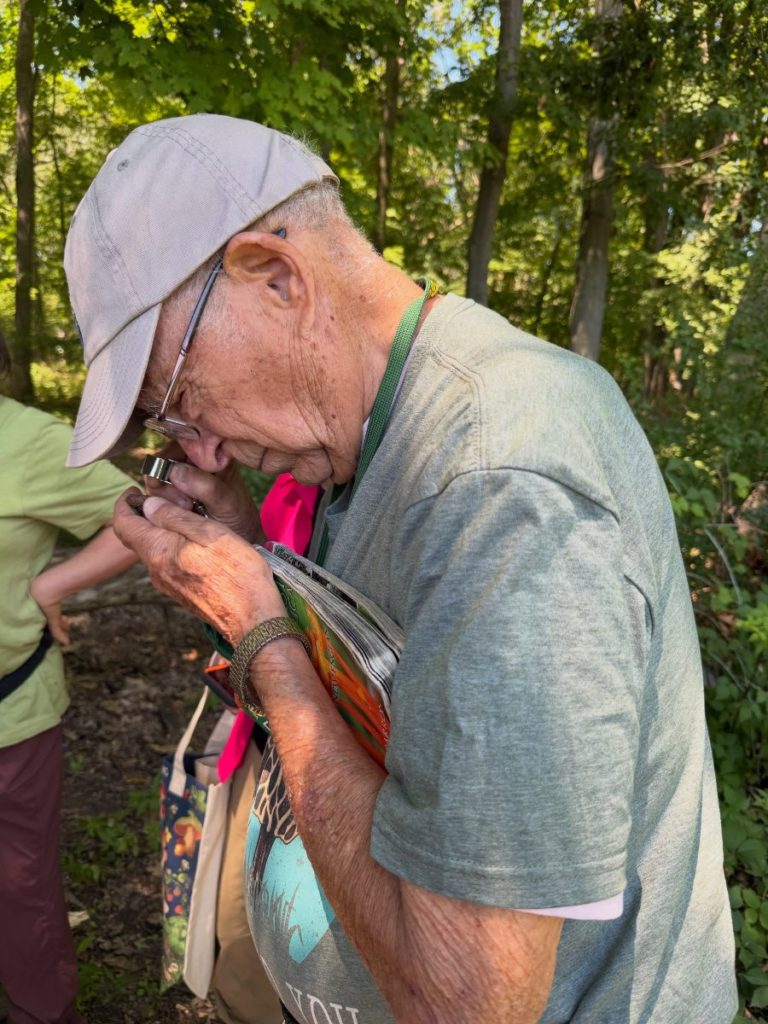 A participant in a cap examining a small specimen with a hand lens on a wooded trail.