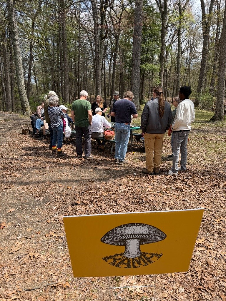 RAMA participants gathered around a picnic table in the woods, with a yellow mushroom-logo sign in the foreground.